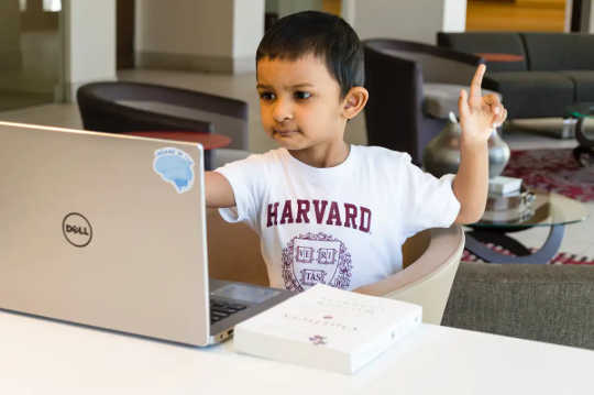 A young boy in a Harvard T-shirt examines a laptop with one finger in the air.