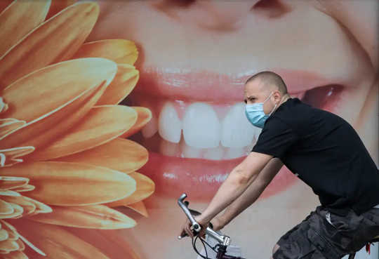 A man wearing a mask cycles past a mural featuring a smiling face in front of a dental office.