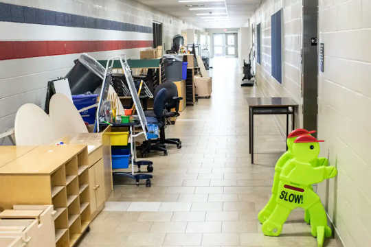 Desks, chairs and shelving units are pushed against the wall of an empty corridor in a school.
