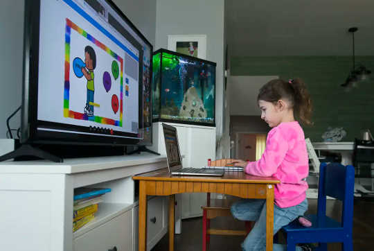 A young girl in a pink sweatshirt sits in front of a laptop and a bigger computer monitor displaying lessons.