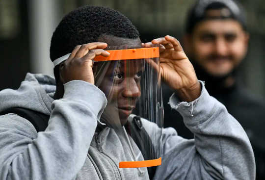 A teenager tries on a face shield with a full face visor as a classmate stands behind him smiling.