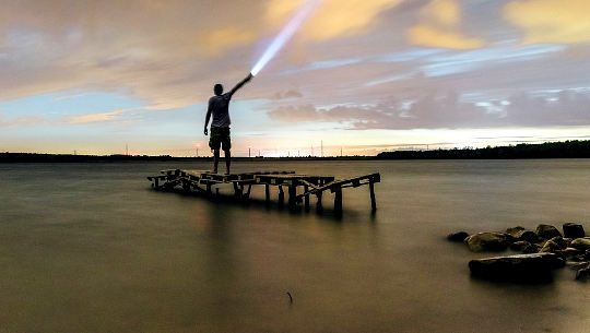 man standing on a dock shining a flashlight into the sky