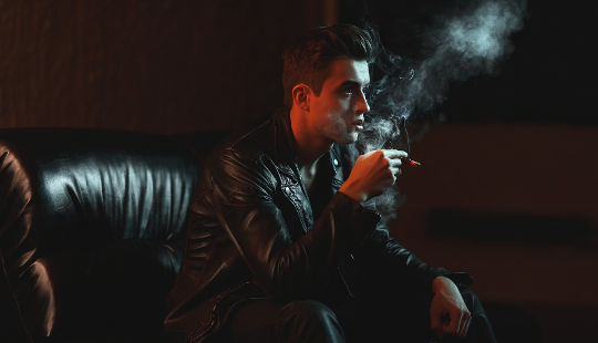 young man sitting in a dark setting smoking