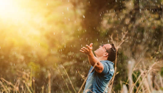 smiling man outside with arms outstretched to the light with drops of rain falling down upon him