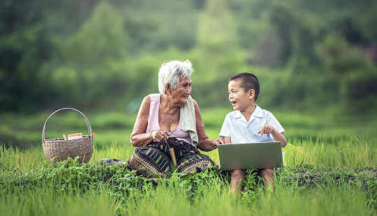 a young child with a laptop speaking with his grandmother sitting outside with a picnic basket