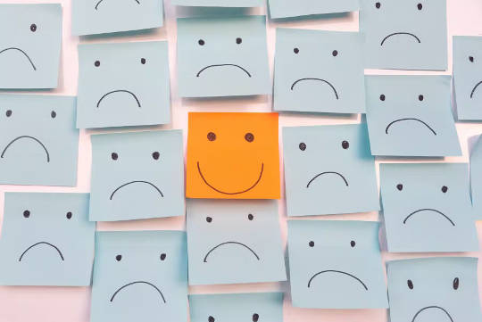 a series of frowning face post it notes on the wall with one bright red smiley-face one