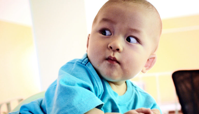 A baby in a blue shirt looks over his shoulder with eyes wide
