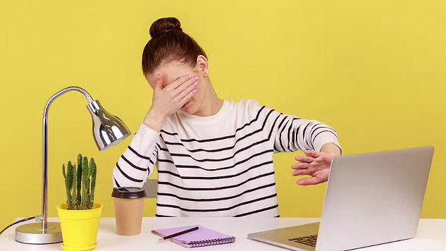 一名年轻女子坐在黄墙前的桌子旁，一只手捂住眼睛，另一只手挡住电脑屏幕，暗示“我不想看这个”。