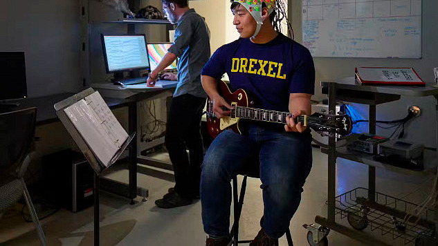 年轻人戴着头盔弹吉他，头盔上覆盖着测量大脑活动的电极
