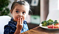 A little girl eats vegetables from a plate while sitting at a table