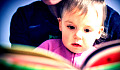 young child on the lap of an adult looking at the pages of a colorful book